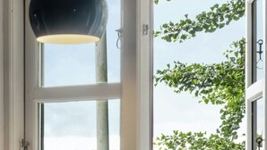 Une fenêtre ouverte donnant sur un champ verdoyant et des buissons, un luminaire suspendu et deux tasses posées sur le rebord de la fenêtre.