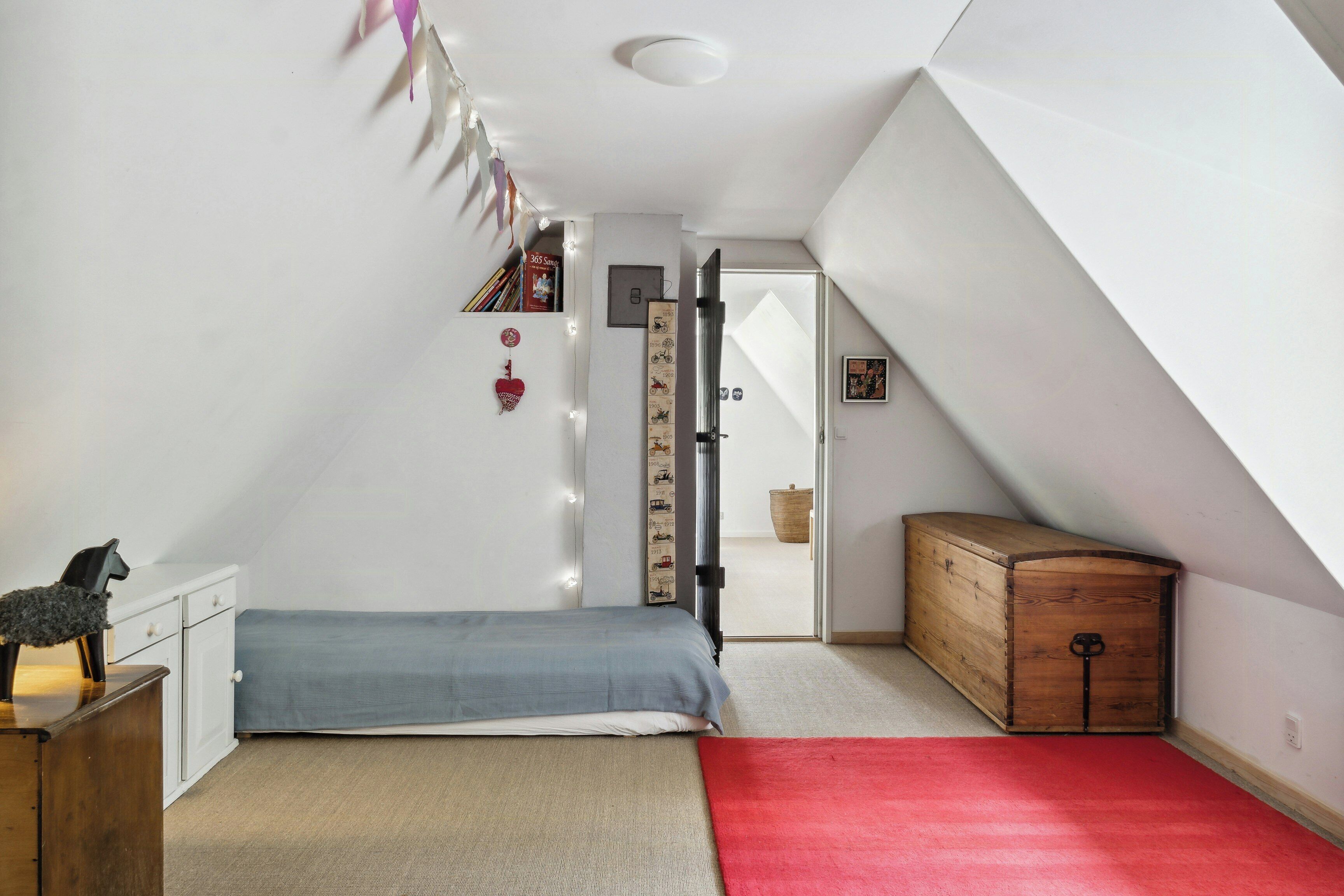 A bedroom with a bed, a dresser, a wooden chest, a red carpet, and a door leading to another room.
