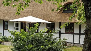 Un bâtiment traditionnel avec un toit de chaume, des murs blancs rehaussés de moulures noires et une fenêtre surmontée d’un parasol blanc.