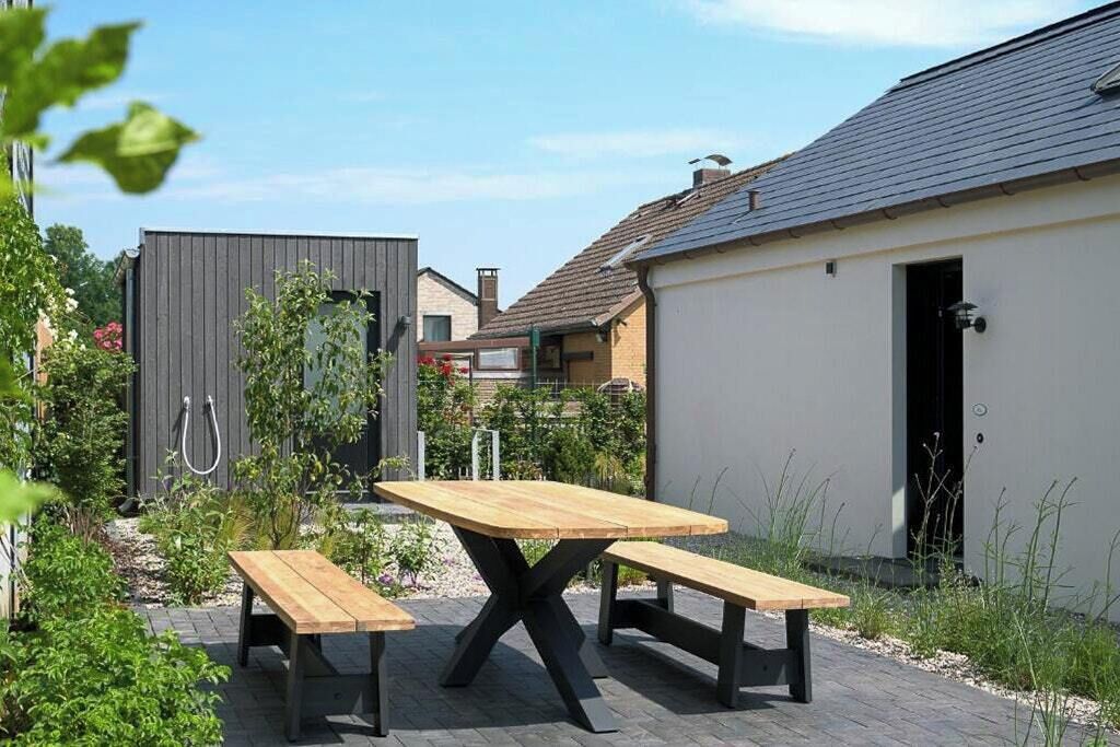 A patio with a wooden table and benches, surrounded by greenery and a building with a grey roof.