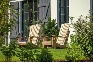 Zwei hölzerne Adirondack-Stühle in einem Garten, im Hintergrund ein Gebäude.