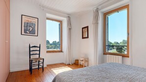 Un dormitori amb llit, una cadira, una taula petita, dues finestres i un quadre amb marc a la paret.
