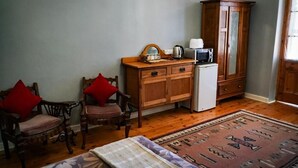 Une pièce comprenant un meuble en bois, un petit réfrigérateur, un four à micro-ondes, deux chaises avec des coussins rouges, un lit recouvert d’une couvre-lit à motifs floraux et un tapis à motifs.