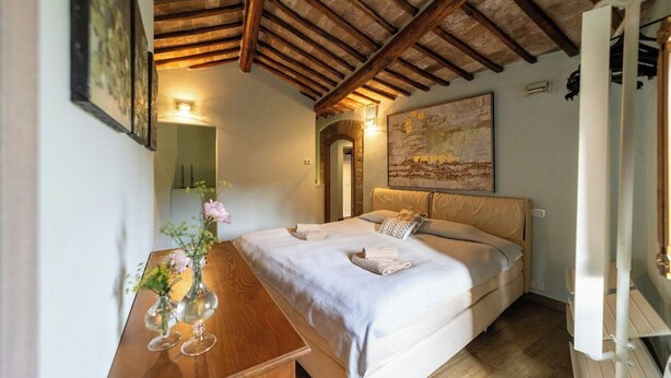 A bedroom with a wooden ceiling, a bed with a beige headboard, a bedside table with a vase, and a painting on the wall. - Villa with heated pool in Toscana (San Quirico)