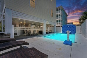 Ein Pool mit blauer Liegefläche, eine überdachte Terrasse mit Gartenmöbeln und ein zweigeschossiges Gebäude mit Balkonen.