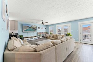 Ein Wohnzimmer mit einem Ecksofa, einem Flachbildfernseher, einem Deckenventilator und Blick ins Freie.