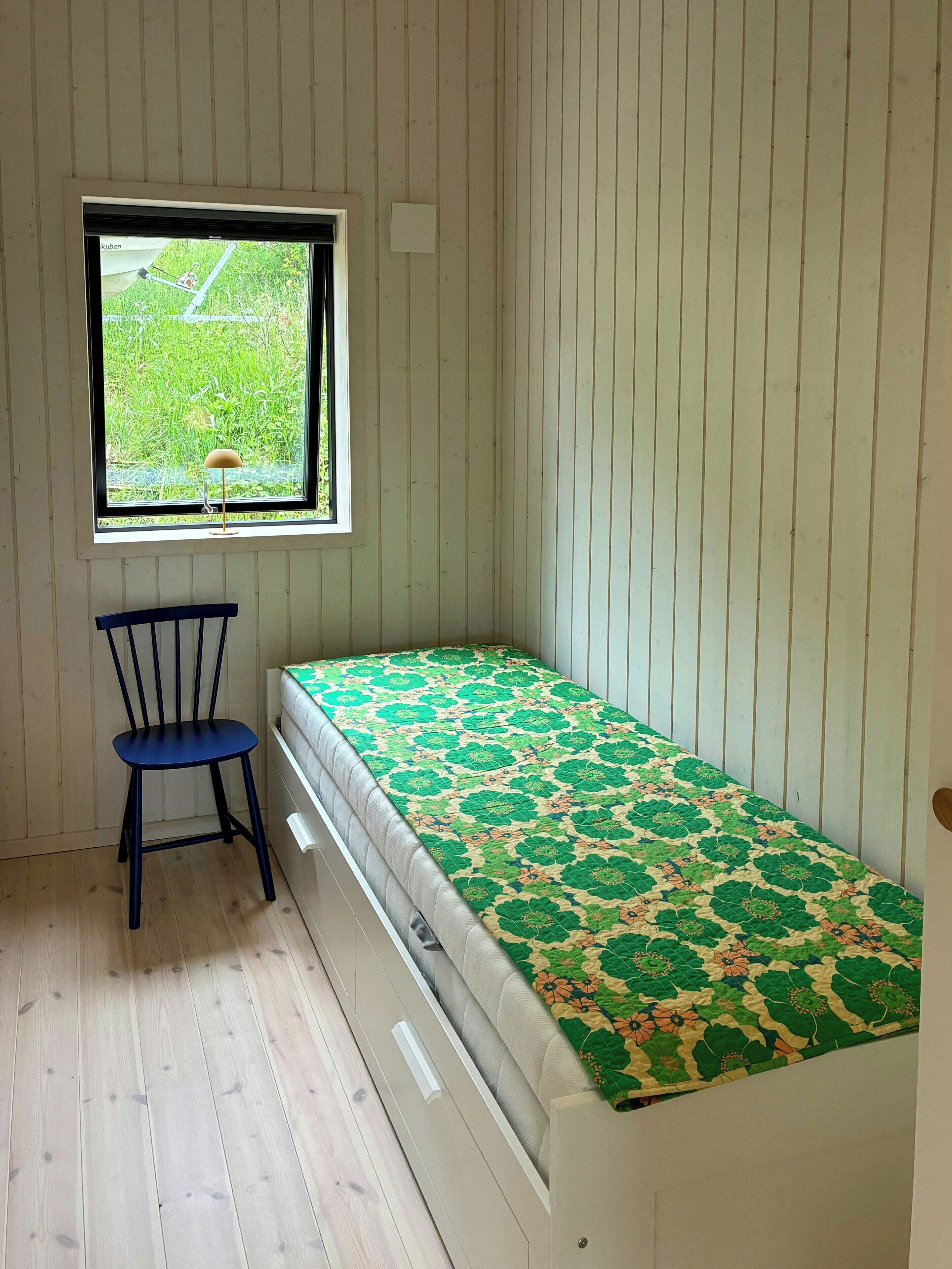 Une petite pièce avec un lit simple, une chaise, une fenêtre et des murs en bois.