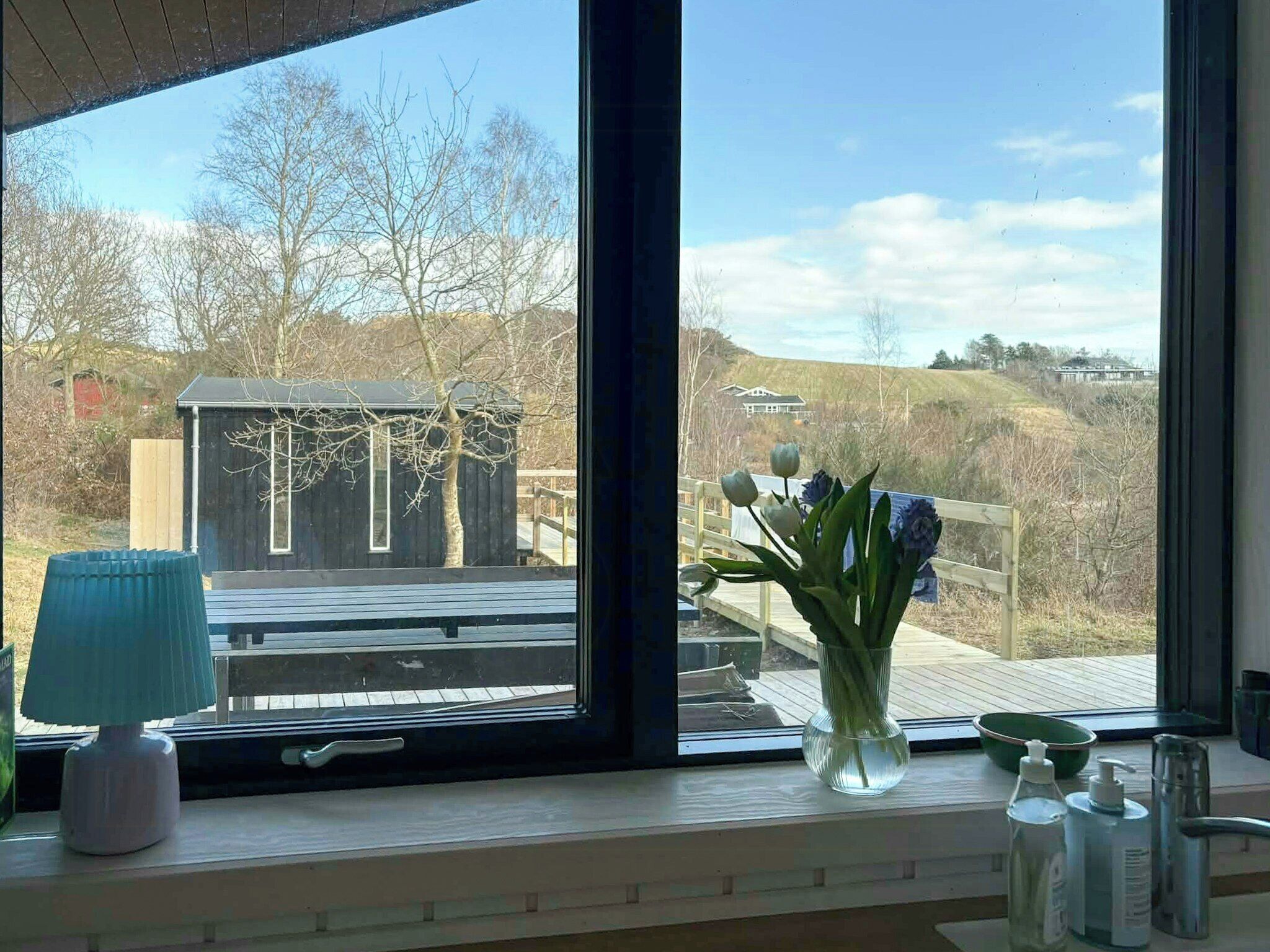 Une pièce dotée d’une grande fenêtre, d’une terrasse en bois, d’un petit abri de jardin et d’un vase rempli de fleurs.