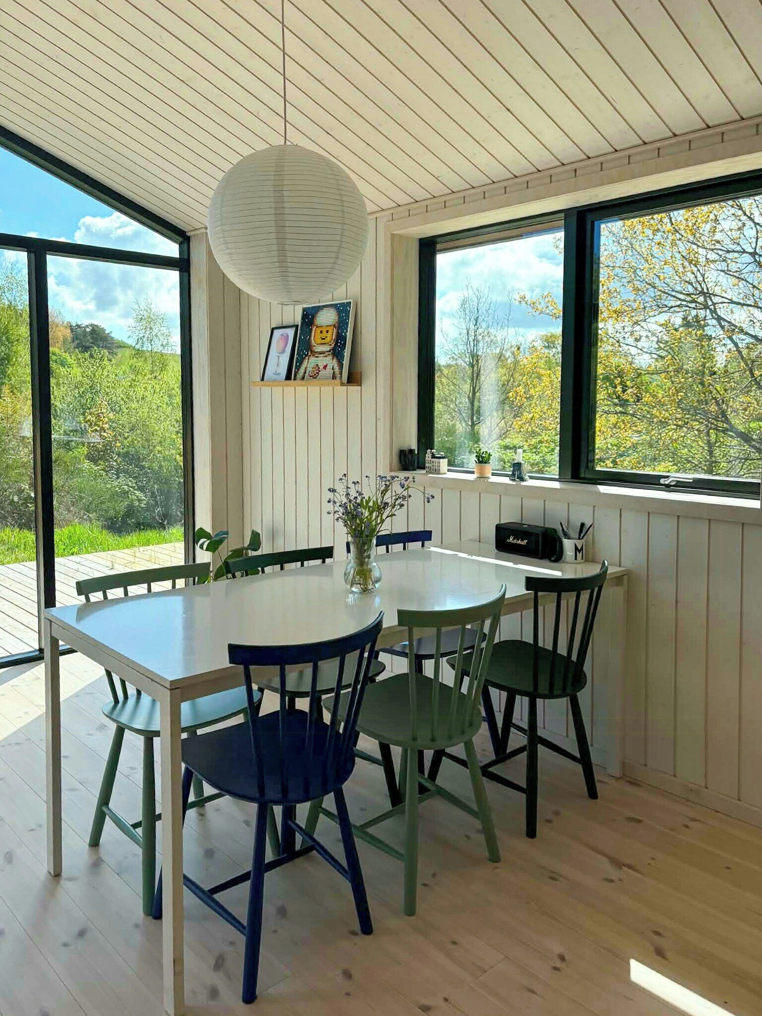 A dining area with a round table, chairs, a hanging light fixture, and large windows offering a view of greenery.