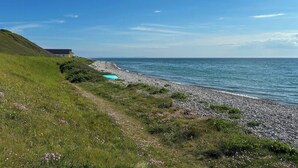 A coastal landscape with a grassy path, a pebble beach, and a building in the distance.