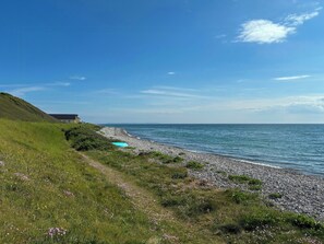 Ett kustlandskap med en gräsklädd stig, en stenstrand och en byggnad i fjärran.