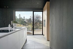 Eine moderne Küche mit einer großen Kochinsel, Schiebetüren zu einer Terrasse und Blick auf Bäume und Himmel.