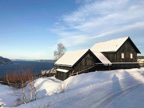 Une cabane en bois, dont le toit est recouvert de neige, située sur une pente enneigée et offrant une vue sur un lac et des montagnes en arrière-plan.