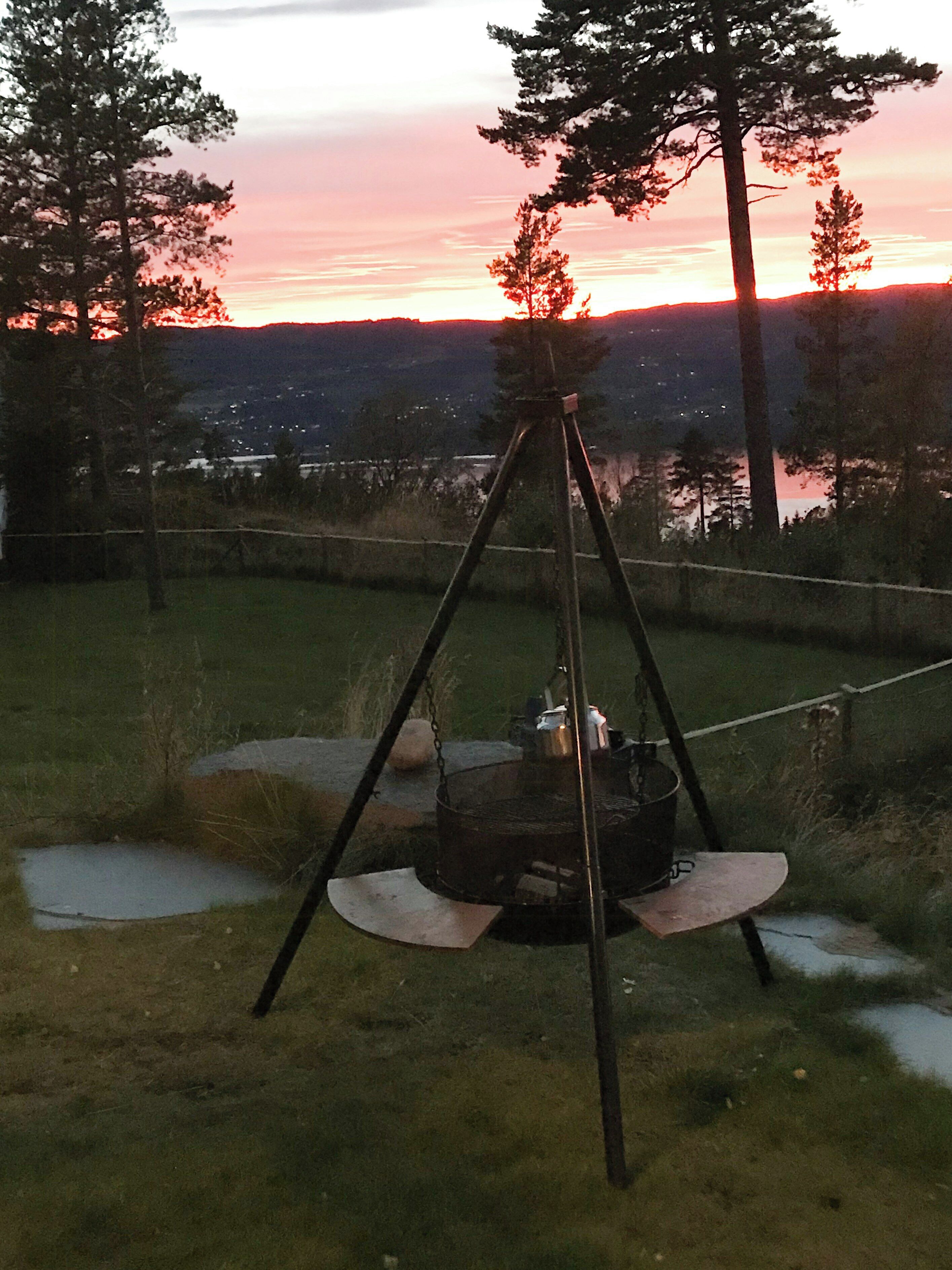 A tripod with a pot on a stand in a grassy area with trees and a distant view of a sunset.