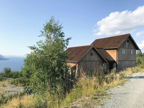 A wooden cabin with a sloped roof, situated on a gravel path with a view of the sea and a tree in the foreground. - Private cabin with stunning views near Oslo (Espa)