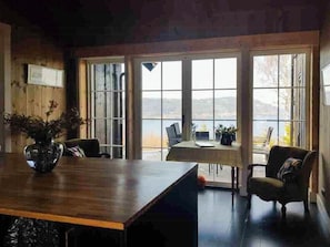 Ein Zimmer mit Holztisch, zwei Stühlen, einer Couch und großen Fenstern mit Blick ins Freie.