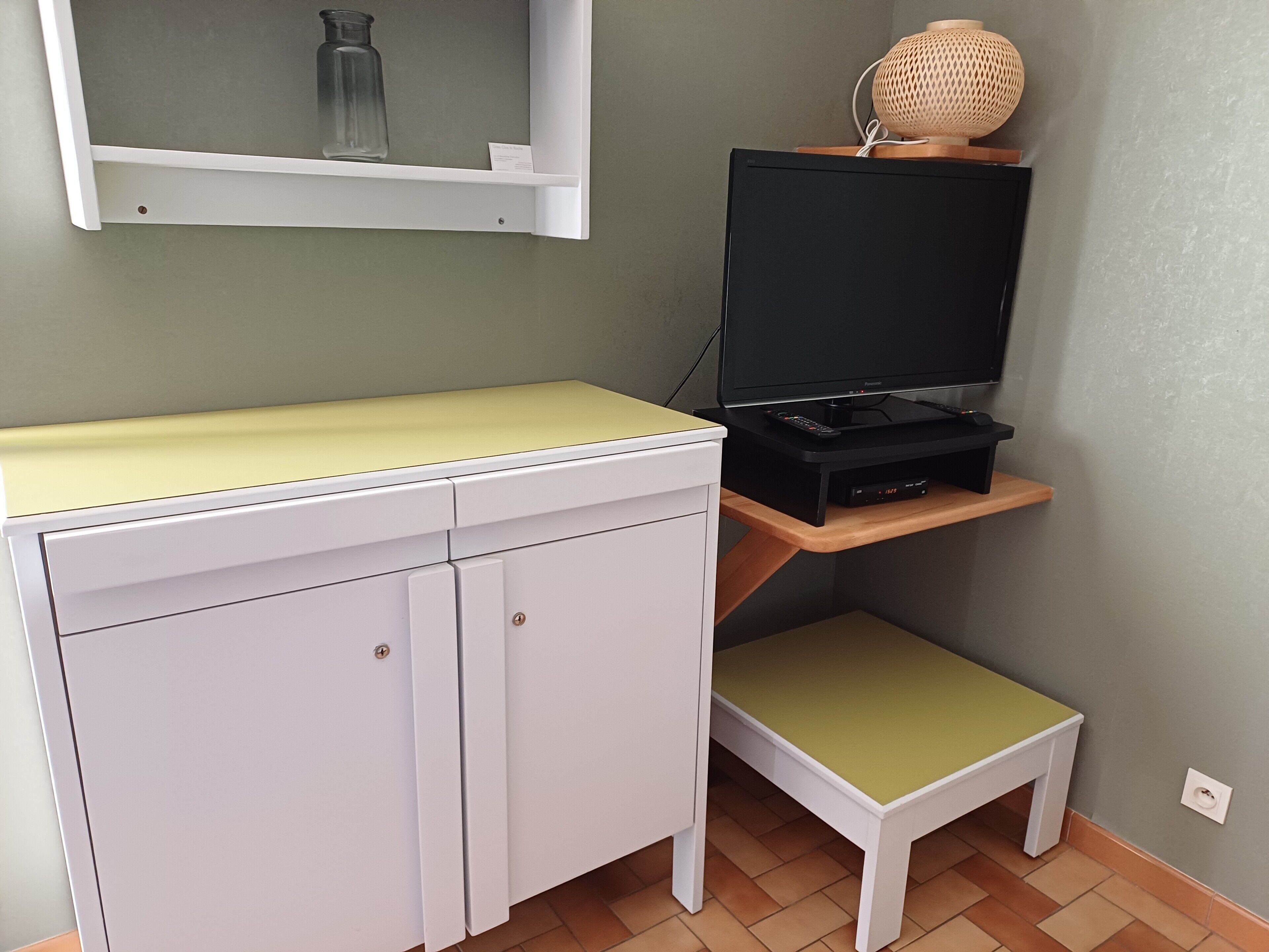 A room with a white desk, a television on a stand, a small shelf with a lamp, and a tiled floor.