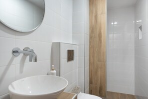 A bathroom with a white sink, a round mirror, a shower, and a wooden panel. - Double 9 Room with Private Balcony, Wi-Fi and Air Conditioning (Ano Symi)