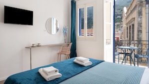 Quarto de hotel com cama, televisão, janela com cortinas, varanda com mesa e cadeiras, e vista para prédios.