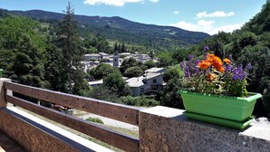 Een balkon met houten hekwerk, uitzicht op een dorp, een groene bloempot met bloemen en een helderblauwe lucht.