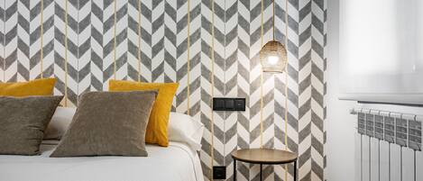 Un dormitorio con pared de diseño en zig zag, una cama con muchos almohadones, una mesita redonda pequeña y una lámpara de pared.