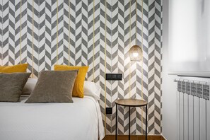 Um quarto com parede em padrão Chevron, uma cama com várias almofadas, uma mesa redonda pequena e um luminário fixado na parede.