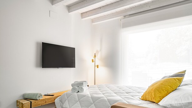 Un dormitorio con cama, televisor montado en la pared, mesita de noche con una toalla enrollada y lámpara de pie.