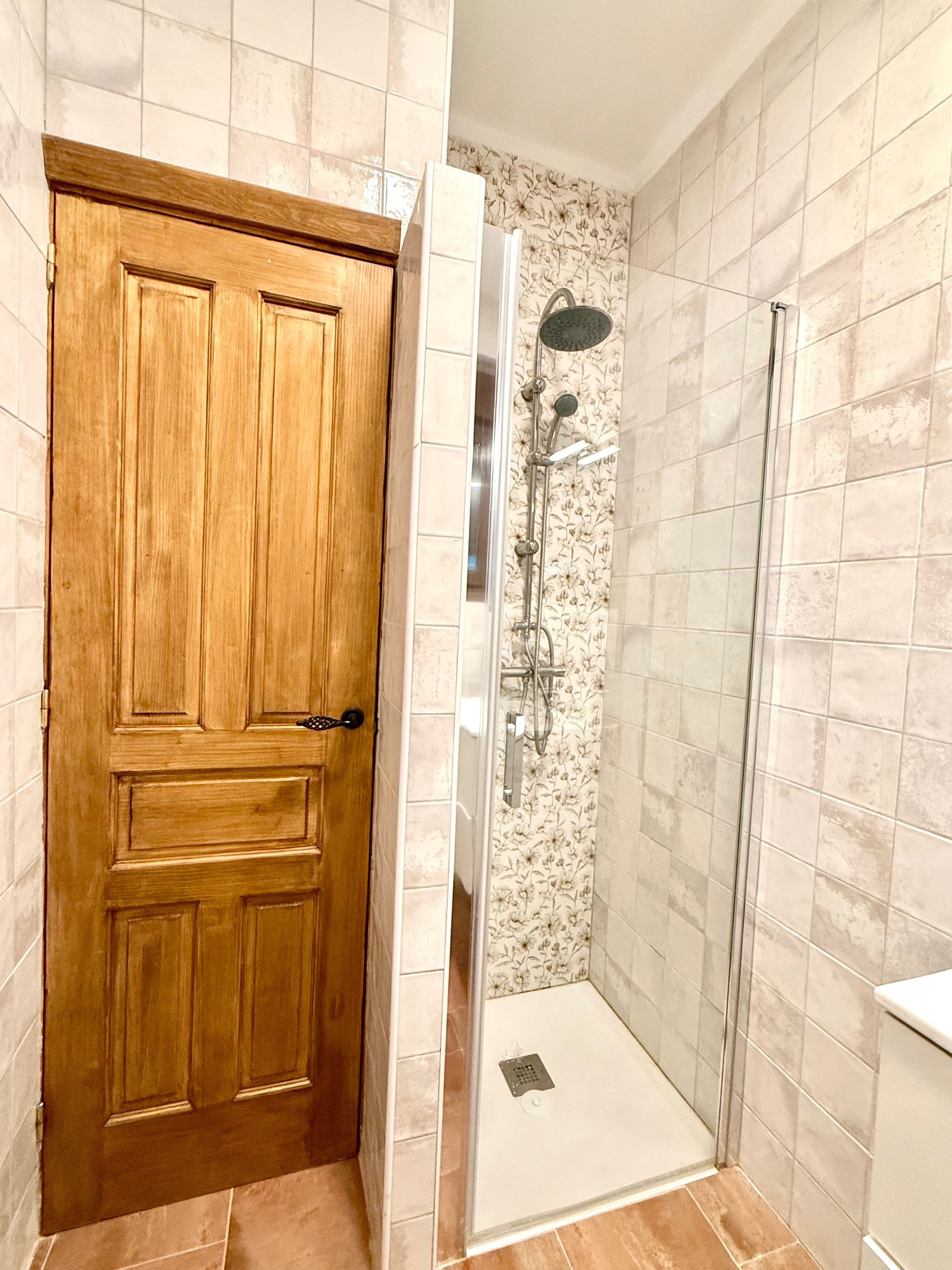 Une porte en bois donne accès à un espace douche, qui est séparé par une porte en verre et équipé d’un pommeau de douche.