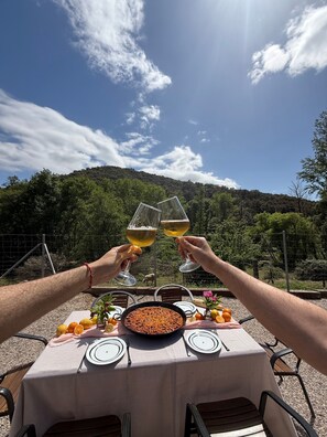 Une table dressée à l’extérieur, avec vue sur une colline boisée, deux verres contenant une boisson jaune et une assiette contenant ce qui semble être un aliment friable.