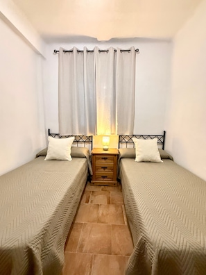 A bedroom with two single beds, a nightstand with a lamp, and a window with curtains.