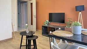 Ein modernes Wohnzimmer mit Flachbildfernseher, einem Esstisch mit Stühlen, einer Topfpflanze und einer Stehlampe.