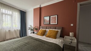 Ein Schlafzimmer mit einem Bett, einem Nachttisch, einer Lampe, einem Fenster mit Vorhängen und zwei gerahmten Bildern an der Wand.
