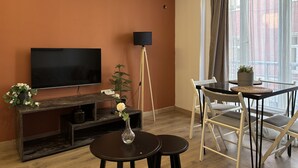 Ein Wohnzimmer mit Flachbildfernseher, Stehlampe, Esstisch mit Stühlen und einem Blumentopf.