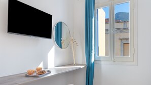 A room with a TV mounted on the wall, a mirror, a window with blue curtains, and a small table with a tray of food. - Studio '11' with Private Balcony, Kitchenette, Wi-Fi and Air Conditioning (Ano Symi)