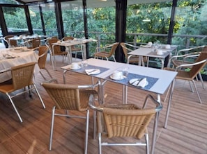An outdoor dining area with tables and wicker chairs, surrounded by glass walls with a view of greenery.