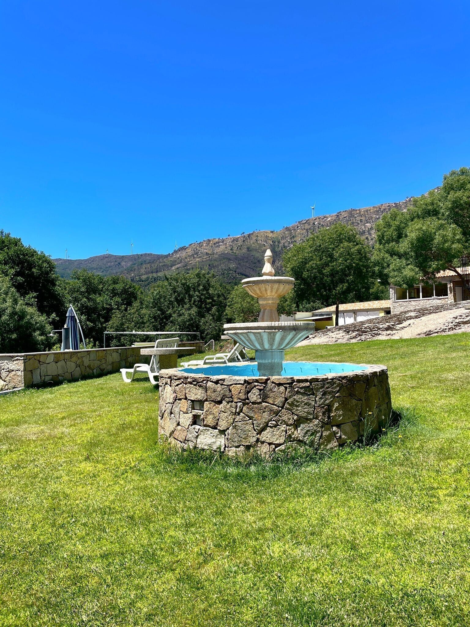 A stone fountain with a pool, surrounded by a stone wall and grass, with a mountainous backdrop.