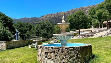 A stone fountain with a pool, surrounded by a stone wall and grass, with a mountainous backdrop.