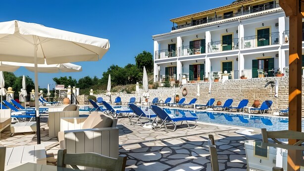 Zona de piscina con sillones y sombrillas, suelo de baldosas de piedra y un edificio con balcones y ventanas.