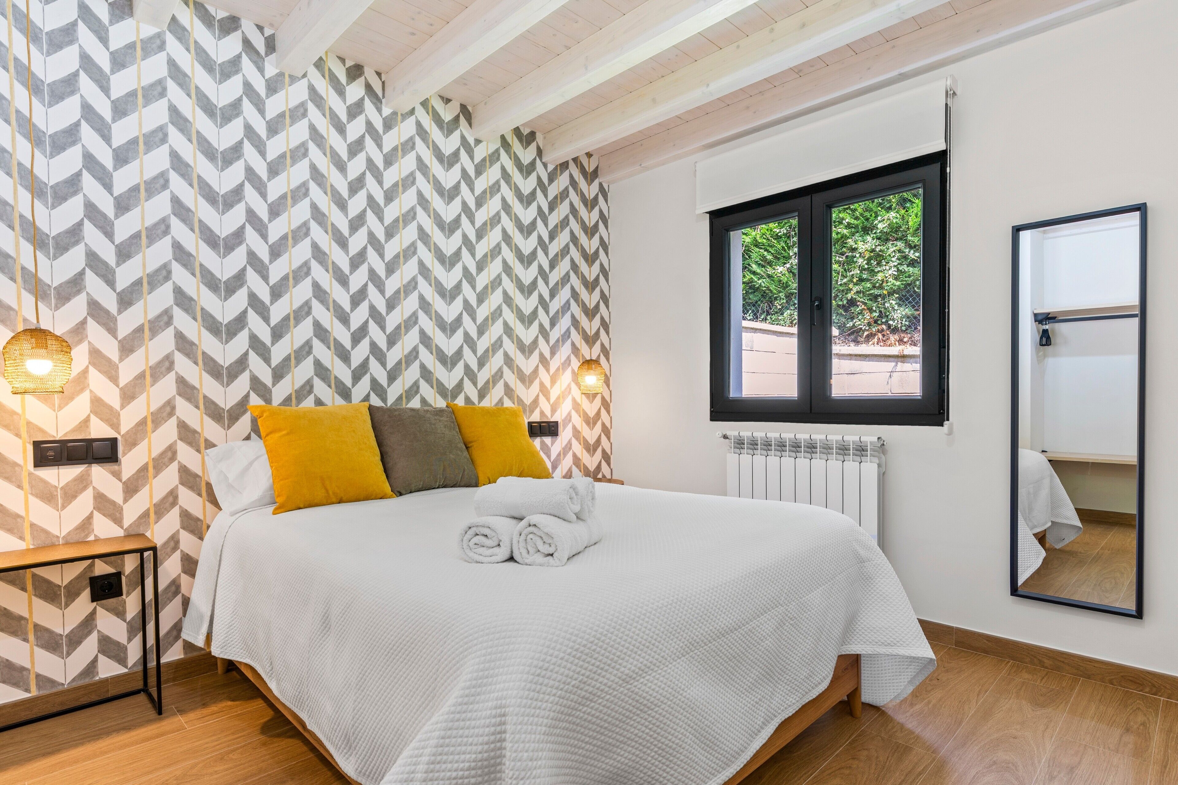A neatly made bed with rolled towels, a bedside lamp, and a window with a view of greenery.