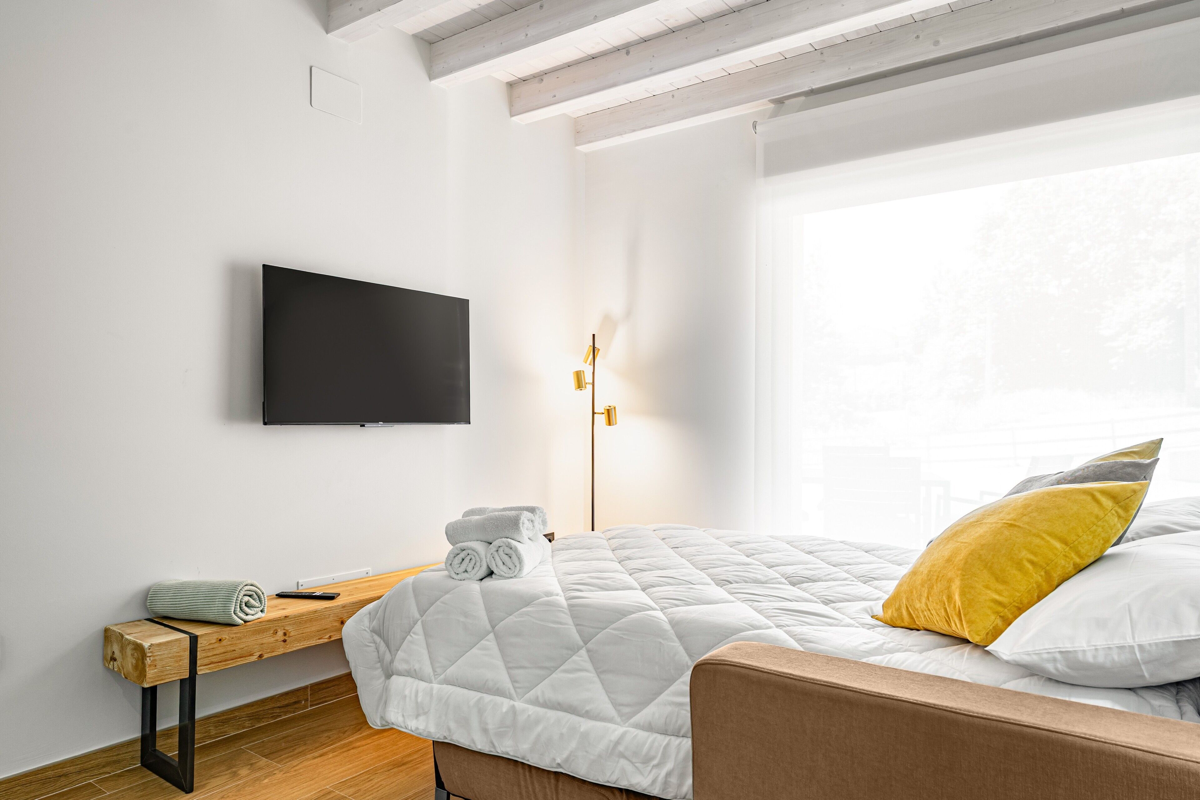 Un dormitorio con cama, televisor montado en la pared, mesita de noche con una toalla enrollada y lámpara de pie.