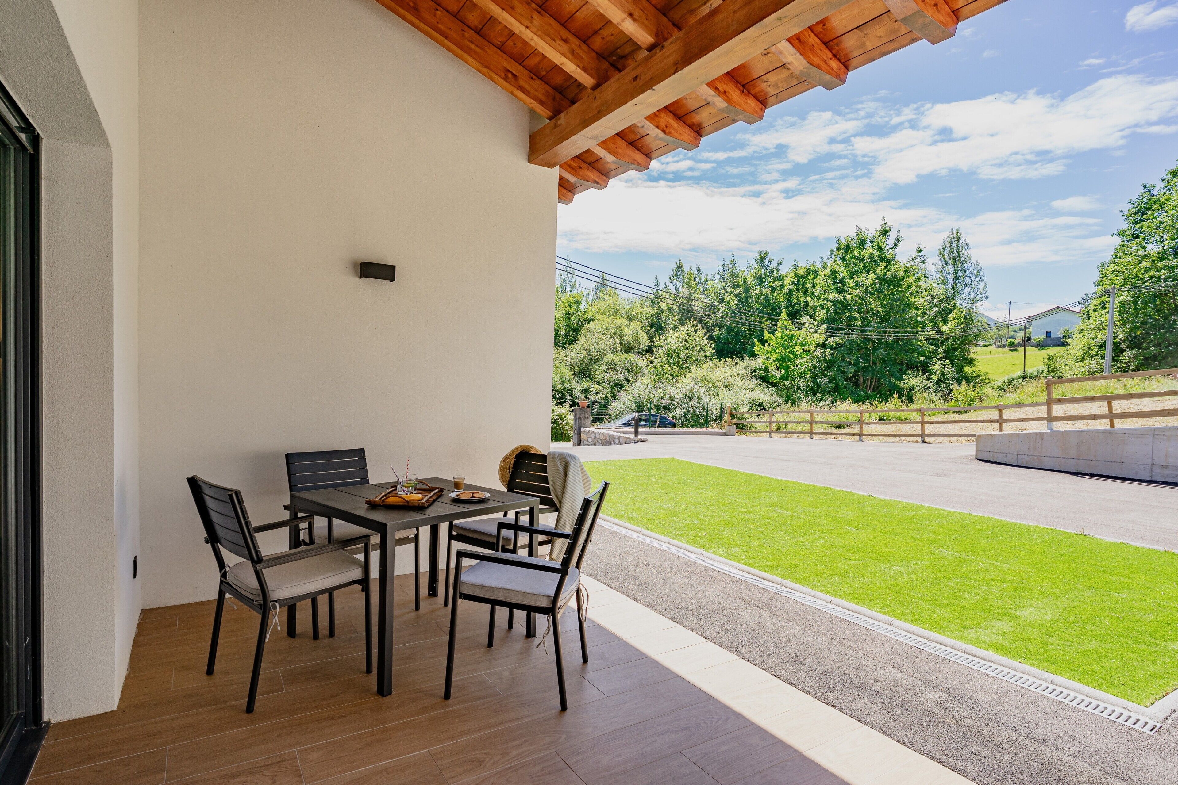 Un patio exterior con una mesa y sillas, techo de madera y vista a un césped verde y árboles.