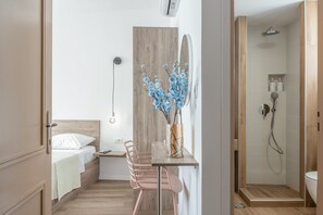 Habitación de hotel pequeña con cama, un escritorio con silla, un florero con flores, zona de ducha y un panel de pared de madera.
