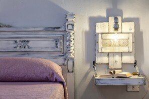 Una cama con cabecera, un aplique en la pared y una estantería con un libro.