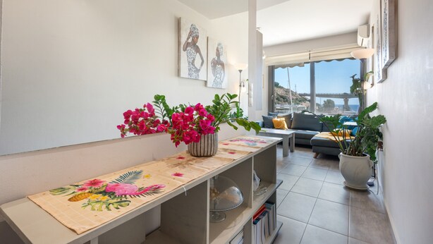 A modern living room with a sofa, a coffee table, a decorative table with a floral table runner, and a large window with a view. - Elva Creta Beach Apartment with Sea View, Wi-Fi, and Air Conditioning (Palaiokastro)