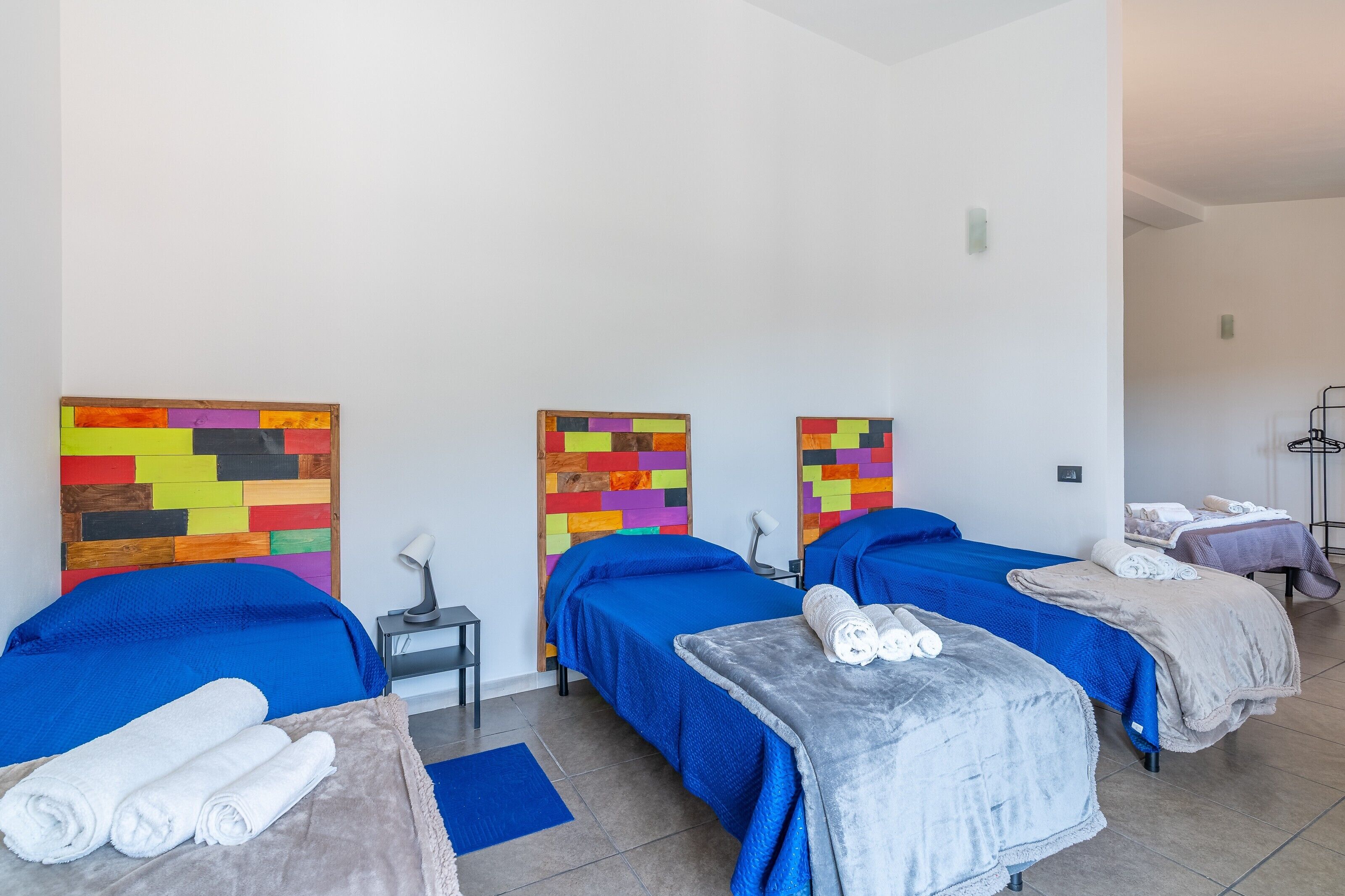 Habitación de hotel con tres camas, cada una con una colcha azul, un cabecero colorido y una mesita con toallas.