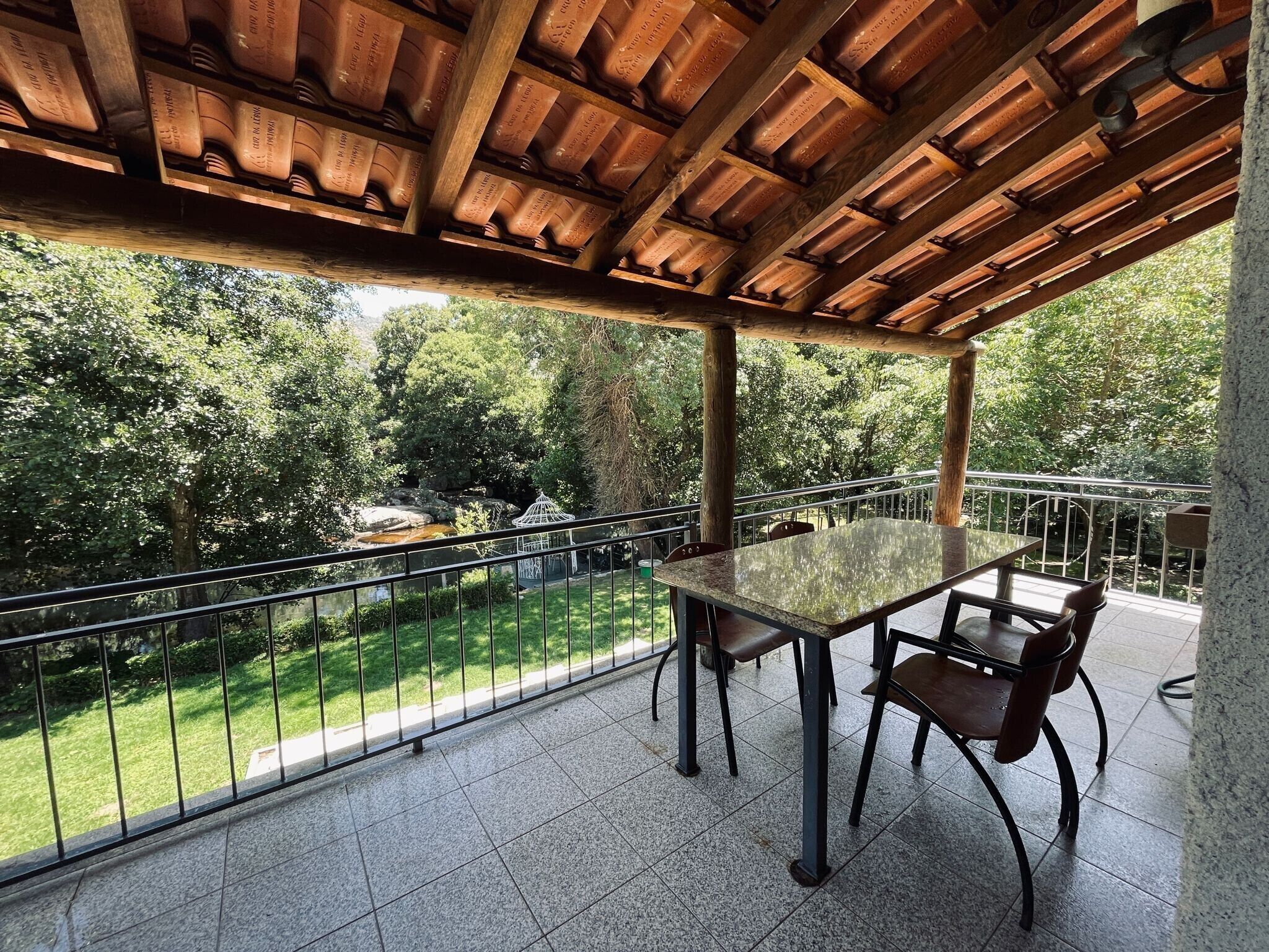 A covered outdoor area with a table and chairs, a railing, and a view of a garden and stream.