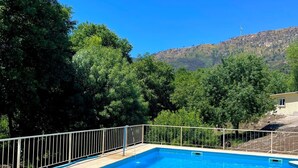 Piscina rectangular con solárium empedrado, rodeada por una valla metálica, con árboles y una colina al fondo.