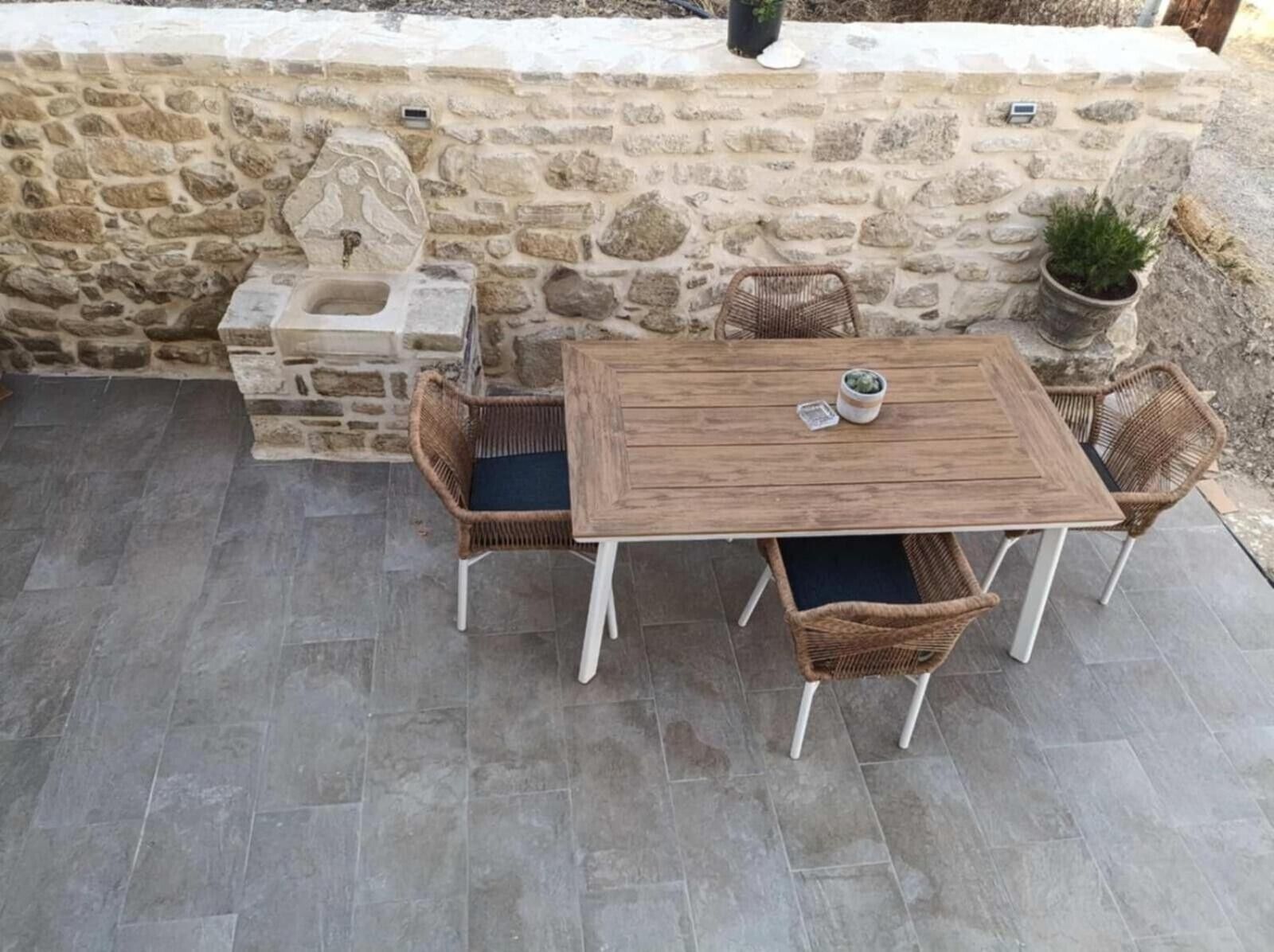 Een stenen muur met een in steen gehouwen reliëf, een houten tafel met vier rieten stoelen, en een bloempot met een plant.