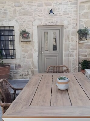 Un edificio de piedra con una puerta marcada '13 1933', una ventana con barrotes, sillas de mimbre y una mesa de madera con una planta en maceta.