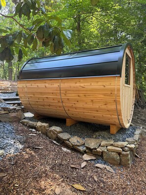 Une structure en bois, en forme de tonneau et surmontée d’un toit noir, entourée d’un mur de pierres et de gravier, située dans une zone boisée.